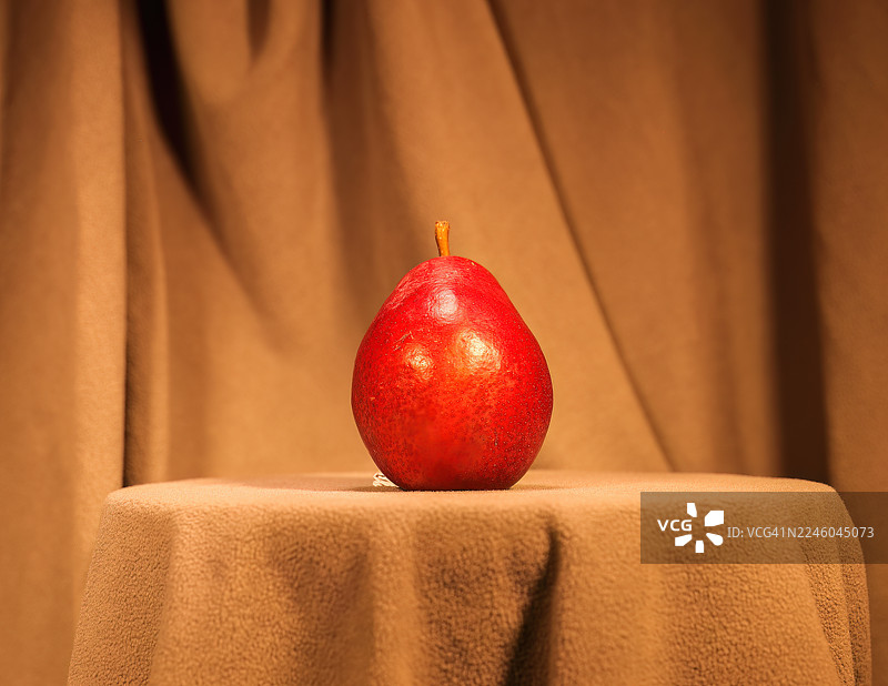 一张红色的梨子放在盖有棕色桌布的桌子上，背景与桌布颜色相配，灯光柔和。图片素材