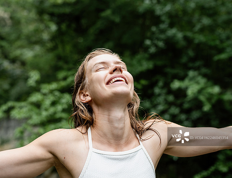 年轻女子在自然环境中，张开双臂，闭着眼睛，仰着脸，享受着雨水，脸上洋溢着喜悦。图片素材