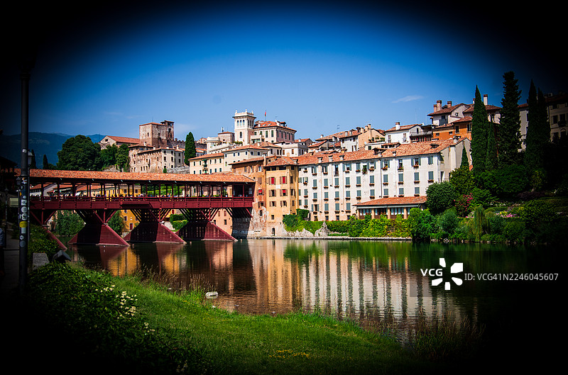 意大利巴萨诺-德尔-格拉帕，布伦塔河上的历史悠久的维奇奥桥，河畔的迷人建筑在晴朗的蓝天下，巴萨诺-德尔-格拉帕，意大利图片素材
