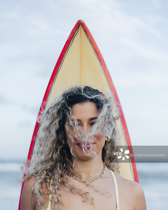 海滩上，一位笑容满面的女士，卷发被风吹拂，手中拿着冲浪板的肖像图片素材