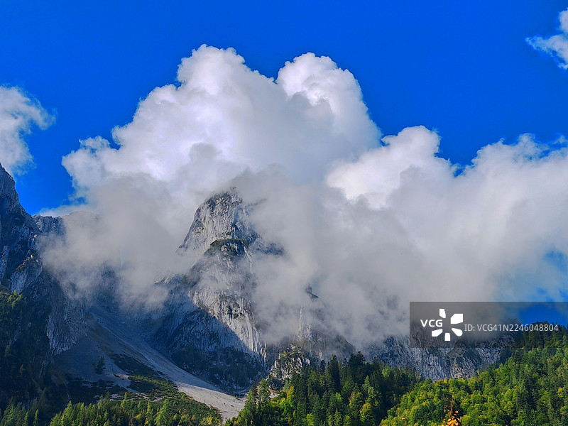 在充满活力的蓝色天空下，雄伟的山峰被戏剧性的云彩笼罩，山脚下是郁郁葱葱的绿色森林图片素材