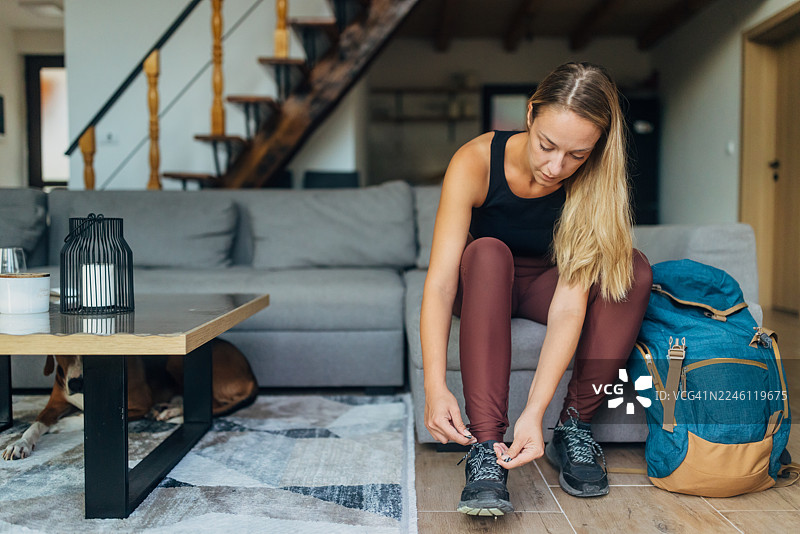 一位女士坐在家里的沙发上，正准备系好登山鞋，为户外探险做准备。她身边放着一个背包，周围是温馨舒适的起居室，装饰温暖。图片素材
