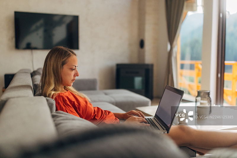 坐在客厅里，透过大窗户和阳台可以看到森林景色，一位专注的女性正在使用笔记本电脑，侧面视角图片素材