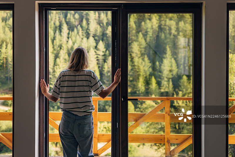 身着休闲装的女子背对镜头，站在现代度假屋的巨大玻璃门前，凝视着窗外郁郁葱葱的绿色山林。图片素材