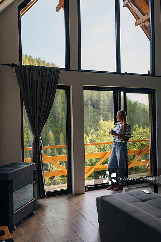 一位宁静的女子手持咖啡杯，若有所思地望着木质阳台栏杆外的广阔阳光下的风景图片素材