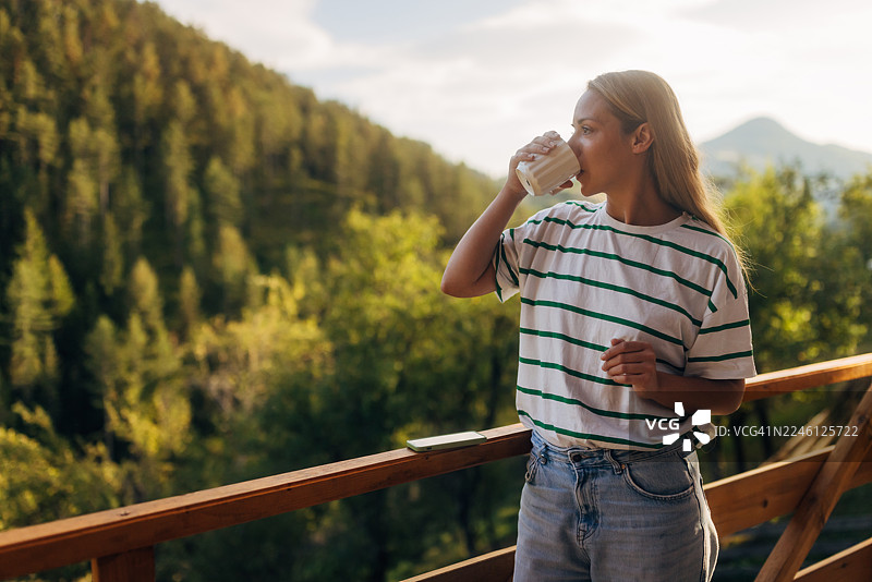 一位美丽的女子在木制露台上，沐浴着自然的晨光，她正啜饮着杯中的饮品，欣赏着翠绿山峦和远方山峰的绝美亮丽景色。图片素材
