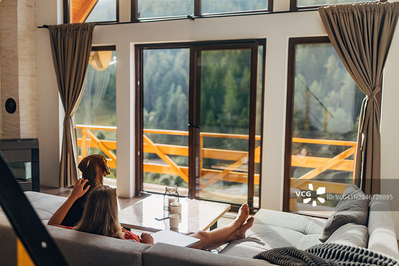 一位女士舒适地躺在沙发上，享受着宁静的早晨，她三色狗在她身边休息。阳光透过大窗户洒进来，在现代客厅里营造出温暖宁静的氛围。图片素材