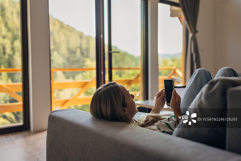 一位无忧无虑的女士躺在客厅的浅灰色沙发上，用智能手机上网和朋友聊天图片素材
