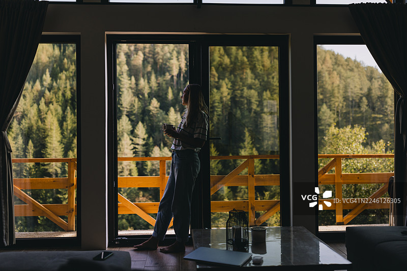 一位穿着休闲服的女子，手持咖啡杯，若有所思地望向一望无际的阳光明媚的风景，她倚靠在木制阳台的栏杆上图片素材