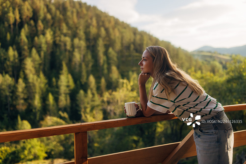 一位女士站在木制露台上，沐浴在清晨的自然光中，她啜饮着杯中的饮品，欣赏着郁郁葱葱的山丘和远处山峰的绝美明亮景色。图片素材