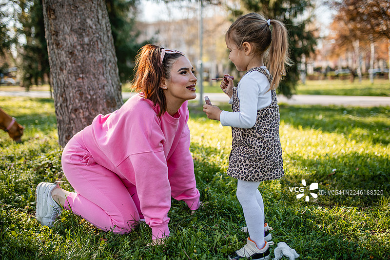 一位漂亮的女士正在公园里和一个可爱的小女孩玩耍，小女孩正在给她化妆。图片素材