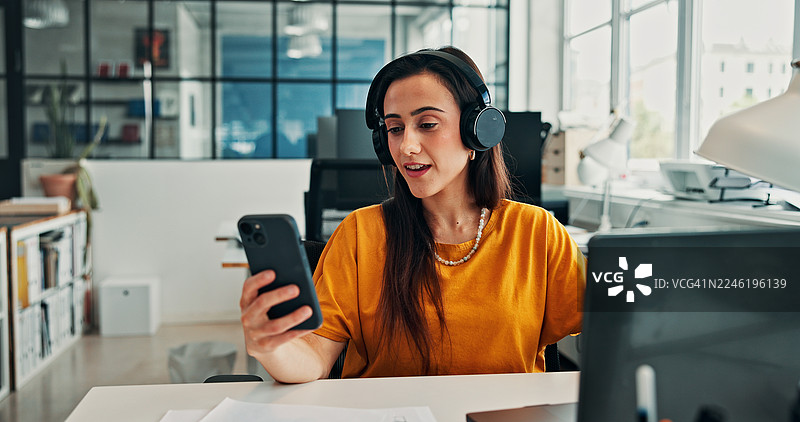女子在办公室戴着耳机、手表，看着手机，背景有音轨，正在工作时听音乐。她面带笑容，正在使用媒体应用程序收听播客或广播订阅的流媒体。图片素材