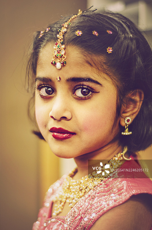 一位身着印度传统珠宝的年轻女孩，包括额饰和发饰，用富有表现力的眼睛望着镜头。她的脸上化着眼线和红唇，突显了文化之美和节日盛装。图片素材