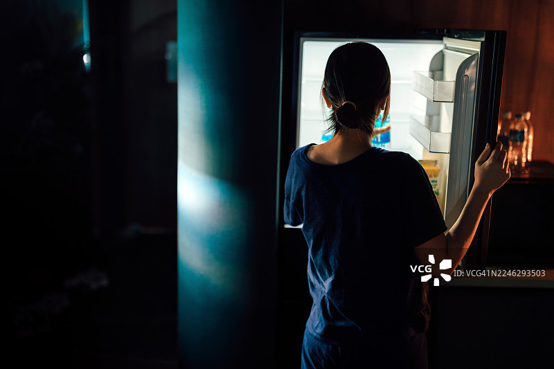年轻亚洲女性的背影，在深夜黑暗的厨房里打开冰箱查看，在家寻找食物。人物与生活方式图片素材