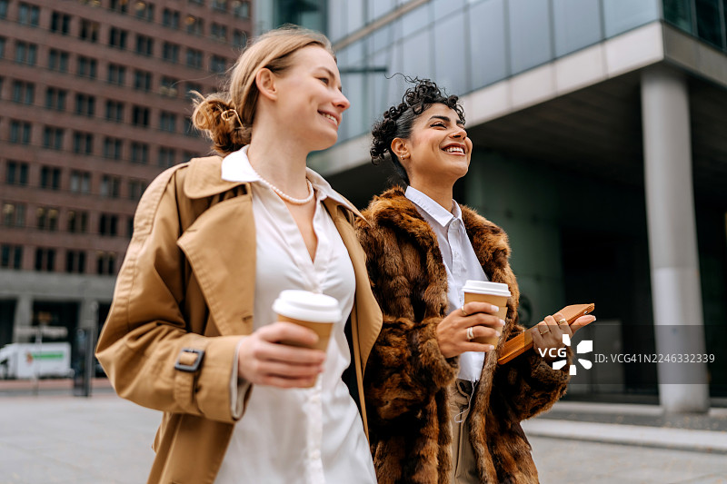 两位衣着光鲜的女士在城市中边走边喝咖啡，并使用平板电脑。图片素材