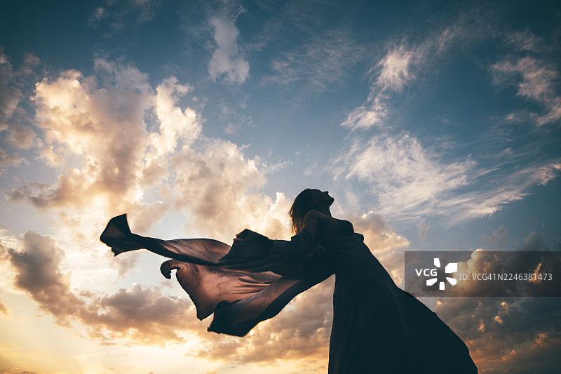 身着飘逸长裙的女子在柔和的云天背景下优雅地漂浮，裙摆随风飞扬，她身体前倾，脸上洋溢着自由与喜悦的笑容。图片素材