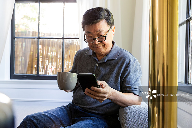 一位年长的亚洲男性在室内放松，一边使用智能手机一边拿着咖啡杯。图片素材