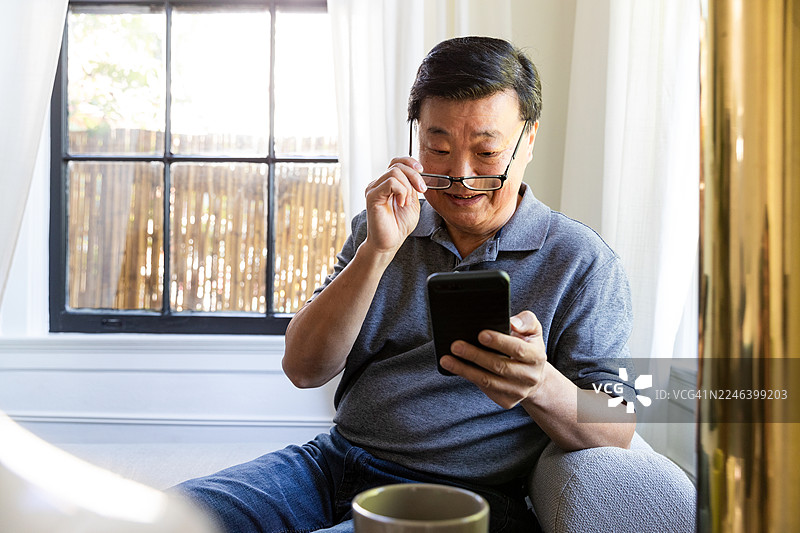 戴眼镜的亚洲男子坐在室内窗户旁，正在使用智能手机。图片素材