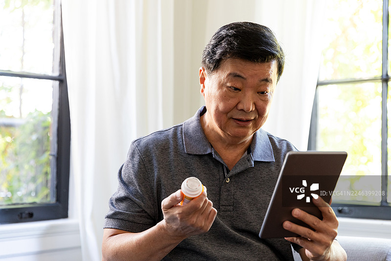 老人室内拿着药瓶使用平板电脑。图片素材