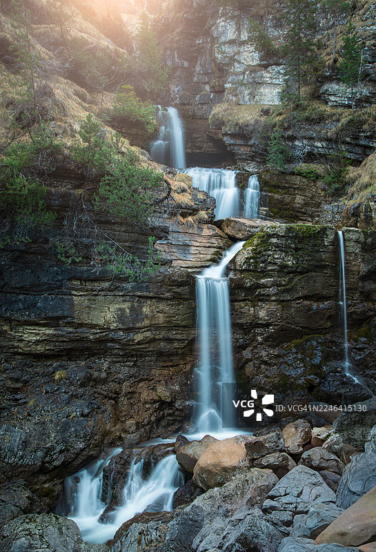 德国福尔尚特，郁郁葱葱的森林中，雄伟的多层瀑布沿着岩石峭壁倾泻而下，阳光穿透树林。图片素材