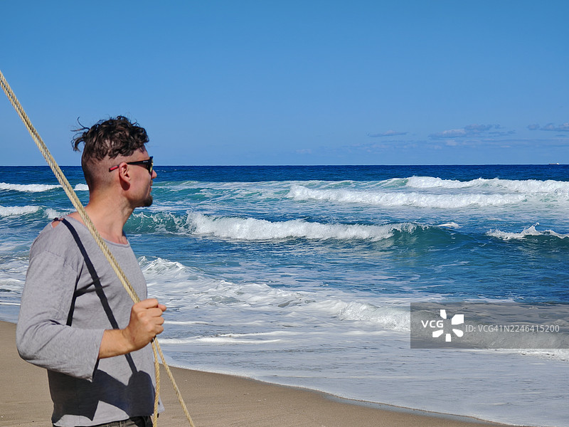 戴着太阳镜的男人站在海滩上，手持绳索，在晴朗的蓝天下眺望海浪，保加利亚图片素材