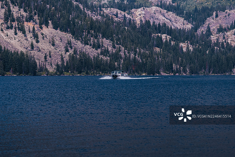 杰克逊，美国，大提顿国家公园，一艘摩托艇在松树林和岩石山脉环绕的广阔湖面上航行，天空晴朗，风景优美图片素材