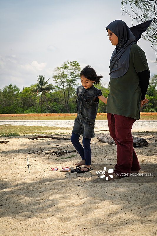 母女俩在阳光明媚的日子里，沿着沙土小径散步，背景是郁郁葱葱的绿植。图片素材