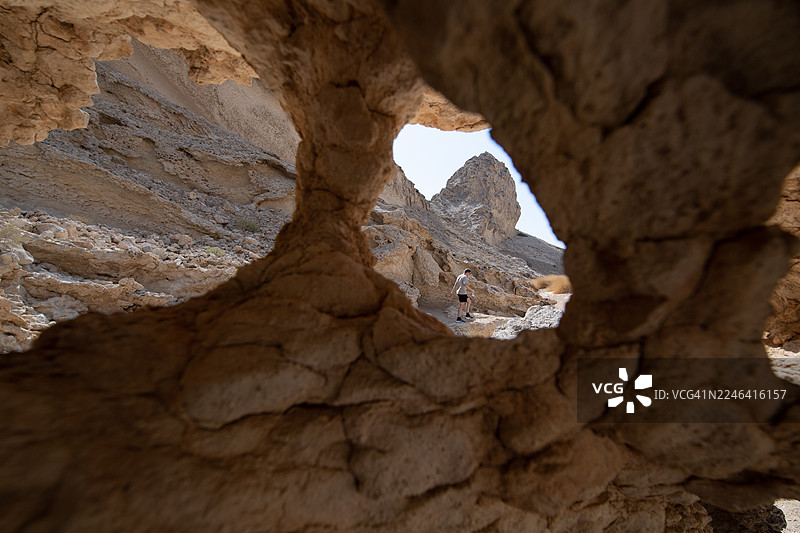 从天然岩石拱门低角度视角看，一个人走在沙漠景观中，背景是山脉，地点是阿联酋艾因的杰贝勒哈菲特。图片素材