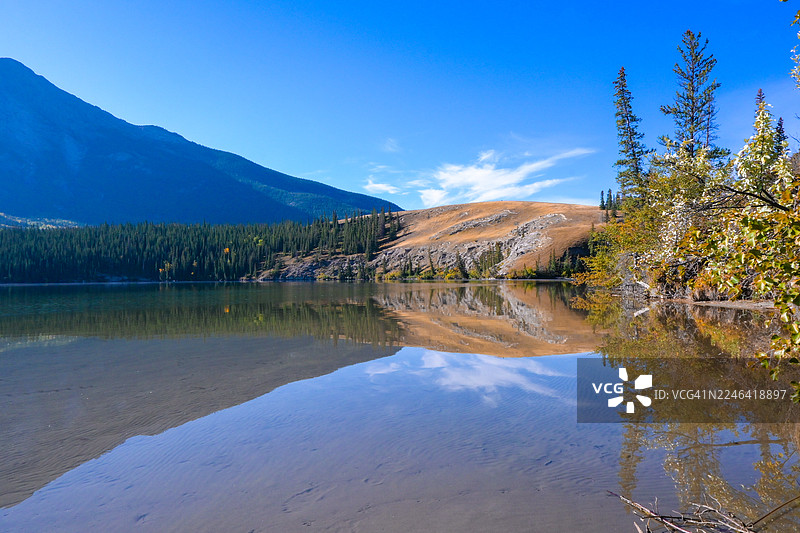 加拿大塔尔伯特湖，晴朗的阳光下，宁静的湖面倒映着远山和湛蓝的天空，稀疏的云彩点缀其间，四周环绕着茂密的常绿森林，一侧是干旱的岩石山坡。图片素材