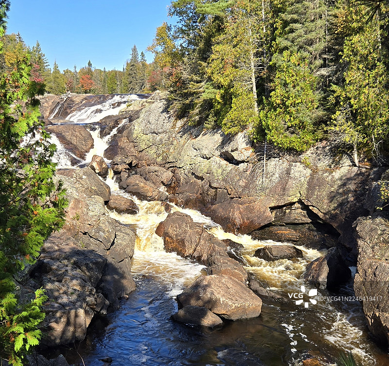 加拿大苏必利尔湖省立公园，雄伟的瀑布从崎岖的岩石上倾泻而下，周围环绕着色彩鲜艳的秋季树木，天空湛蓝。图片素材