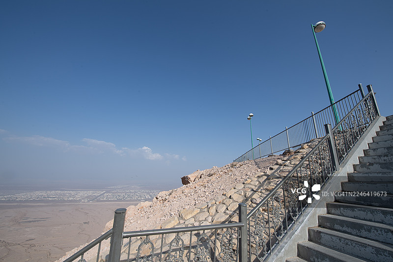 仰视角度拍摄，混凝土台阶配有金属栏杆，沿着岩石山坡向上延伸，通往晴朗的蓝天之下，俯瞰着远处的城市景观。地点：阿联酋艾恩，杰贝勒哈菲特。图片素材