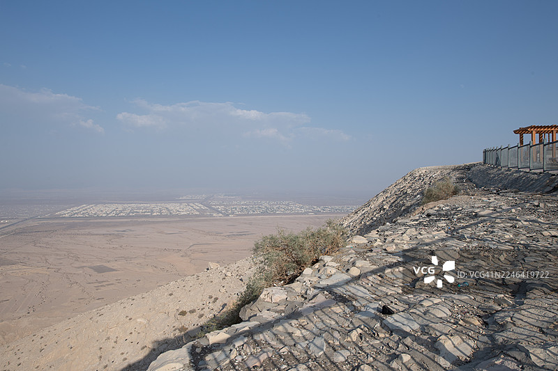 广阔的沙漠景观，远处的城镇沐浴在晴朗的蓝天下，从岩石山坡望去，阿联酋艾因，杰贝勒哈菲特图片素材