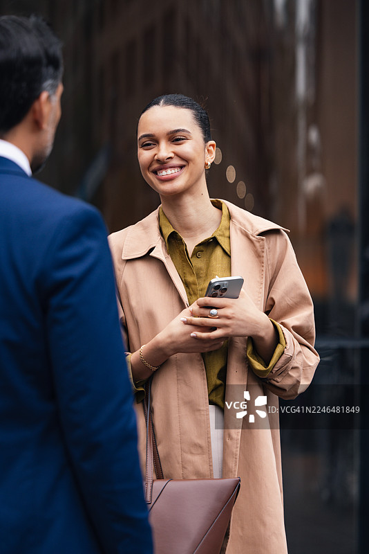 忙碌的街道上，一位微笑着的女商人正与同事交流，她手中拿着手机和手提包，看起来像是在进行一次户外会面。图片素材