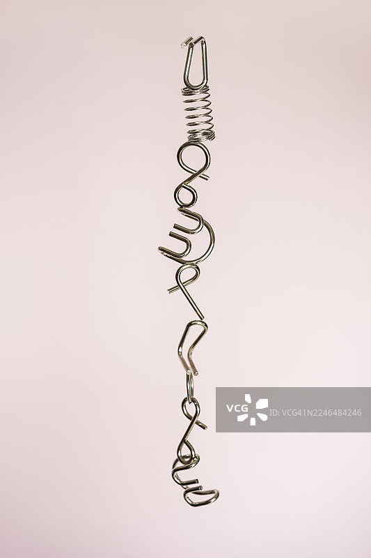 极简抽象构图，一根扭曲的金属弹簧和线状结构垂直悬挂在柔和的粉色背景上，营造出平衡、脆弱和工业优雅的感觉。图片素材