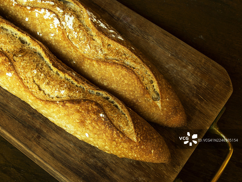 特写镜头：一块带有黄铜手柄的木板上放着两个新鲜、外皮酥脆的面包。图片素材