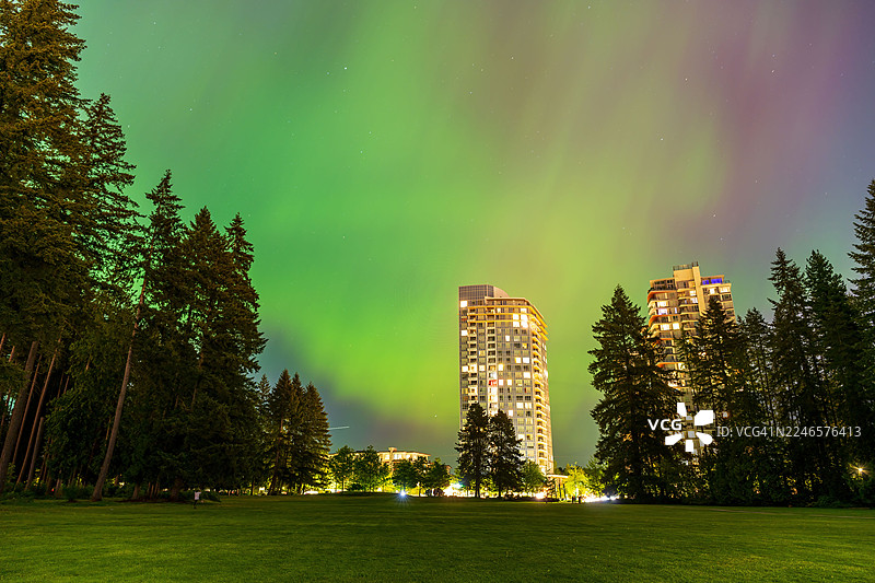 绚烂的北极光照亮了城市夜空，其上空是高耸的建筑和茂密的绿树。图片素材