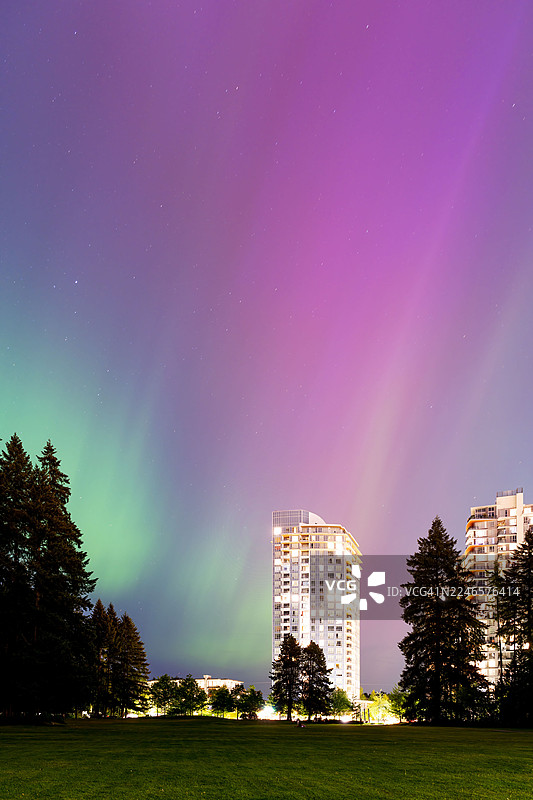 夜空下灯火辉煌的建筑低角度视图，可见绿色和紫色的北极光和星星图片素材