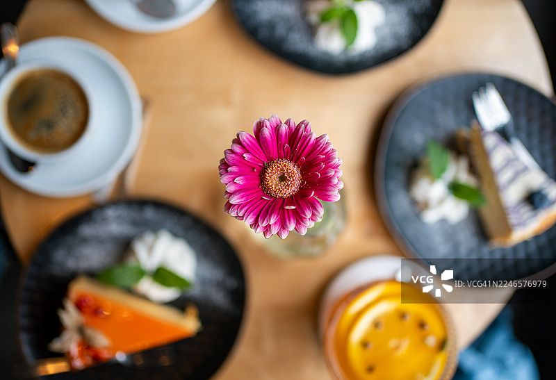 俯视视角，桌上有一朵鲜艳的粉色非洲菊，旁边摆放着咖啡、蛋糕和饮品，营造出温馨的咖啡馆氛围图片素材