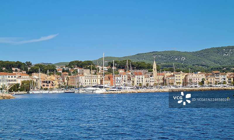 法国卡西斯，晴朗蓝天下，海港停泊着满载船只，色彩缤纷的建筑和教堂塔楼，背景是郁郁葱葱的绿色山丘，风景如画的滨海小镇景色。图片素材