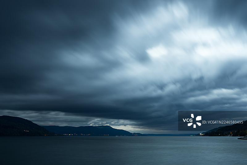 挪威埃兹沃尔市明内松德的远山和城市灯光下的湖泊或海洋，在阴沉多云的天空下，采用戏剧性的长曝光拍摄。图片素材