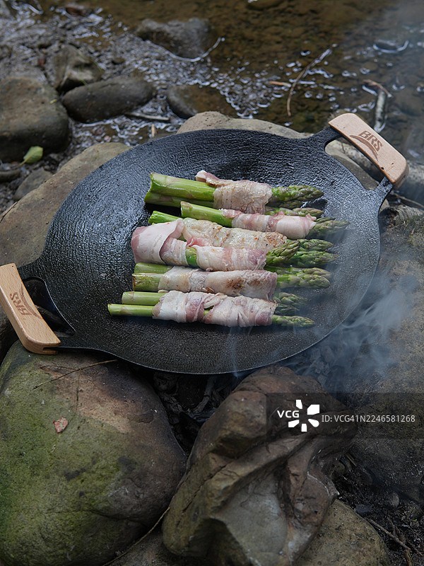 培根卷芦笋在河边的铸铁锅中用明火烧烤，展示户外烹饪和露营美食图片素材