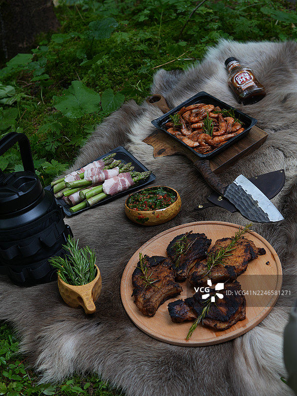 俯瞰视角，森林里的户外盛宴，有烤肉、虾、培根卷芦笋和新鲜香草，摆放在毛皮地毯上，看起来十分美味。图片素材