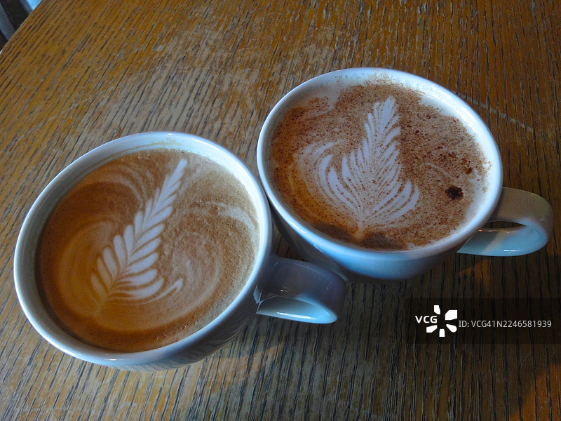 俯视角度拍摄的木桌上两杯带有拉花艺术的美味卡布奇诺咖啡，非常适合悠闲的咖啡时光图片素材