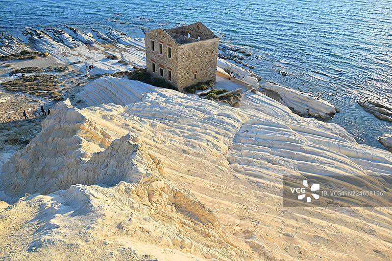 从高处俯瞰，阳光明媚下，一座古老的石砌建筑坐落在崎岖的海岸线上，旁边是碧蓝的大海。地点：意大利阿格里真托，蓬塔比安卡海滩。图片素材