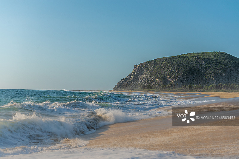 海浪拍打着沙滩，背景是郁郁葱葱的绿色山坡，天空湛蓝，地点是墨西哥埃尔佩斯卡德罗。图片素材