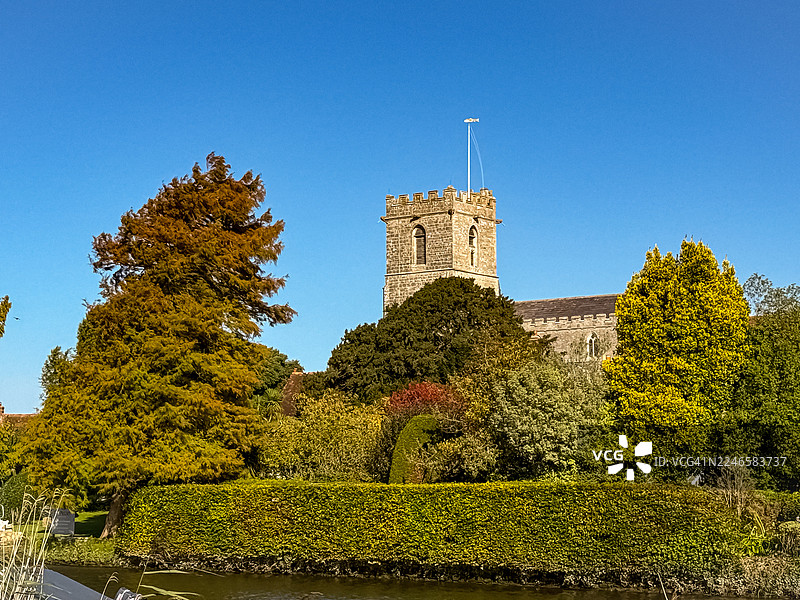 阳光明媚的晴天，在湛蓝的天空下，历史悠久的教堂塔楼高耸于郁郁葱葱的绿树和树篱之上，位于英国瓦勒姆的阿恩自然保护区。图片素材