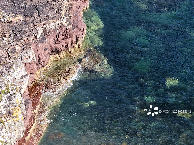 阳光明媚的日子里，高角度俯瞰崎岖的岩石构造与清澈湛蓝的海水相遇图片素材