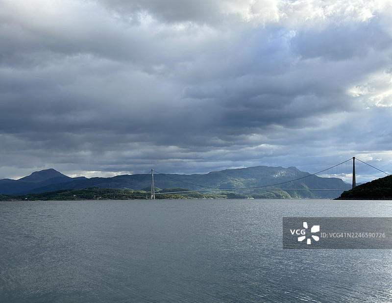 广阔大海上的戏剧性多云天空，远处的山脉和一座悬索桥图片素材