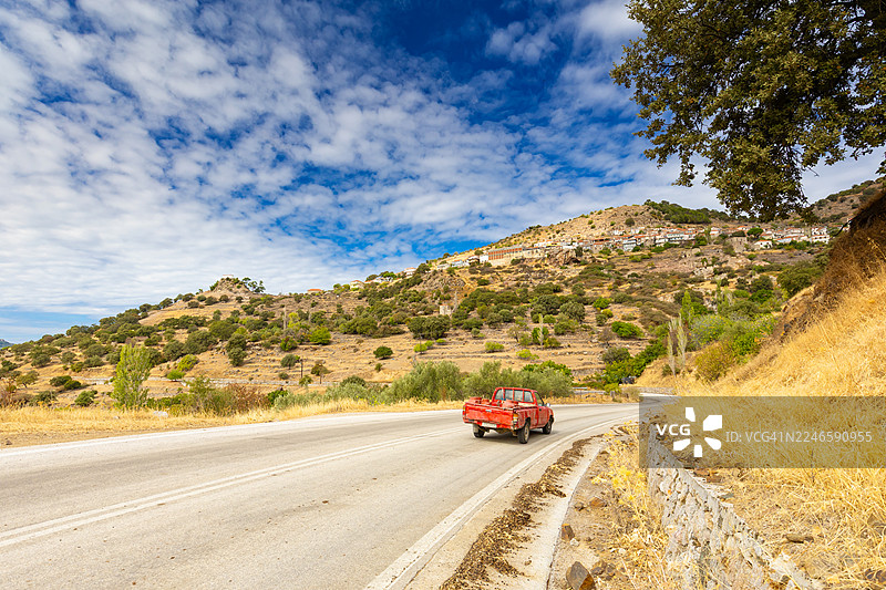 一辆红色皮卡车在希腊莱斯博斯岛的干旱山区蜿蜒的道路上行驶，远处有一个村庄，天空湛蓝。图片素材