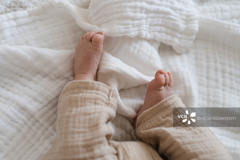 俯视角度，一个婴儿舒适地躺在家中白色的床上，露出小小的脚丫和柔软的衣物。图片素材